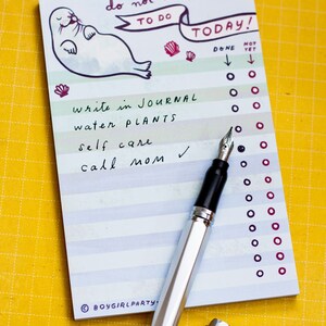 May include: A notepad with the text "do not want to do TODAY!" and a seal illustration. The list includes "write in journal", "water plants", "self care", and "call mom". A silver fountain pen with a black grip and cap is on the notepad.