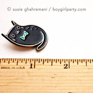 May include: Black enamel pin of a cat wearing a turquoise bow tie. The pin is about 1 inch wide and is shown next to a ruler.
