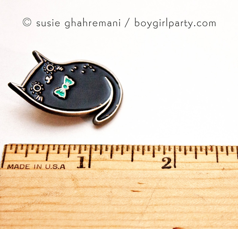 May include: Black enamel pin of a cat wearing a turquoise bow tie. The pin is about 1 inch wide and is shown next to a ruler.