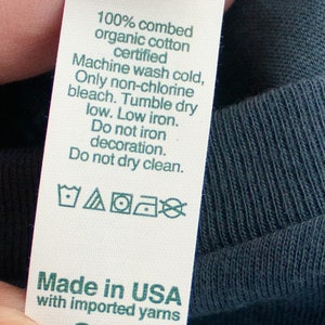 May include: A white care label with green text and symbols. The label reads "100% combed organic cotton certified", "Machine wash cold, Only non-chlorine bleach. Tumble dry low. Low iron. Do not iron decoration. Do not dry clean.", and "Made in USA with imported yarns".