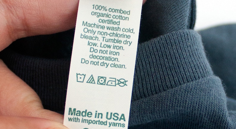 May include: A white care label with green text and symbols. The label reads "100% combed organic cotton certified", "Machine wash cold, Only non-chlorine bleach. Tumble dry low. Low iron. Do not iron decoration. Do not dry clean.", and "Made in USA with imported yarns".