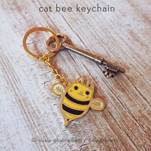 May include: A gold keychain featuring a cartoon cat bee with black and yellow stripes. The keychain also includes a gold key.