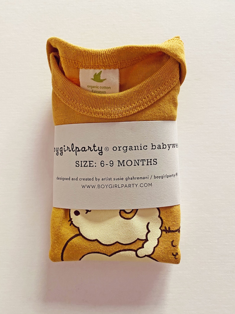 May include: A mustard yellow organic cotton baby onesie with a cute cartoon illustration of a cat. The onesie is folded and has a label that reads "boygirlparty organic babywear" and "SIZE: 6-9 MONTHS".