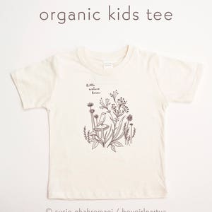 May include: A white organic cotton kids tee shirt with a brown illustration of a bird sitting on a mushroom in a field of flowers. The text "little nature lover" is printed on the shirt.