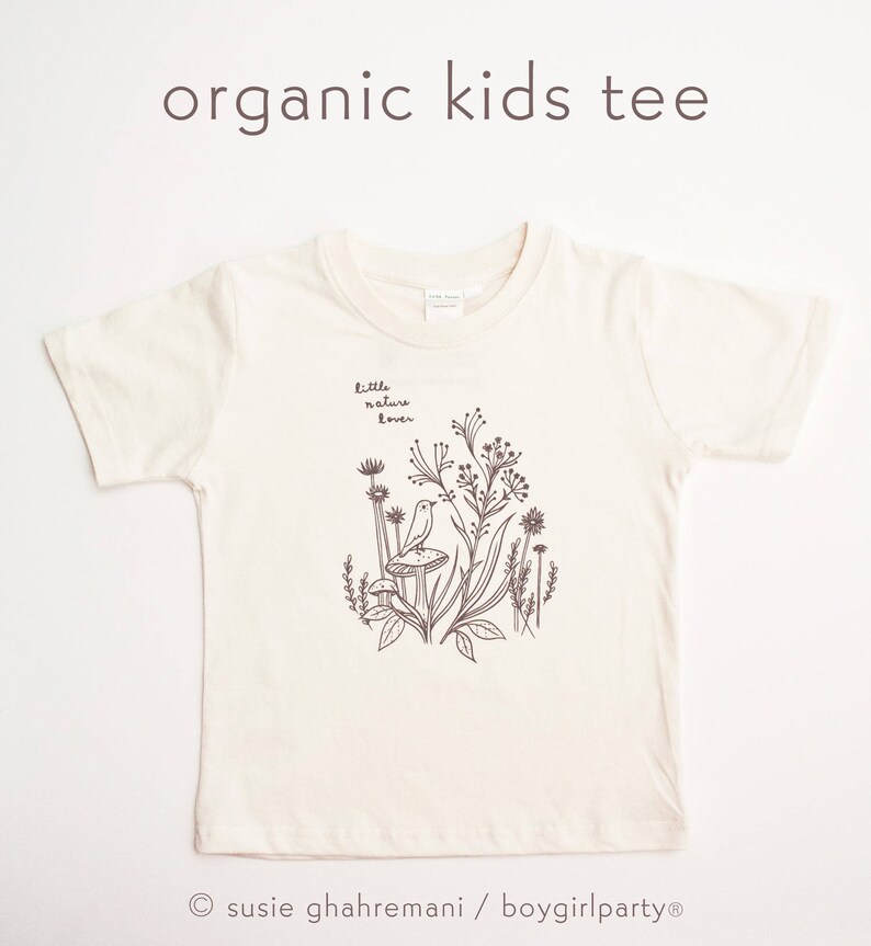 May include: A white organic cotton kids tee shirt with a brown illustration of a bird sitting on a mushroom in a field of flowers. The text "little nature lover" is printed on the shirt.