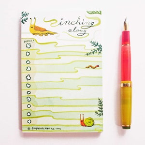May include: A green and white to-do list notepad with a snail and slug illustration. The notepad has the text "inching along" and "DONE?" with a checklist. The notepad is on a white background with a red and yellow pen.