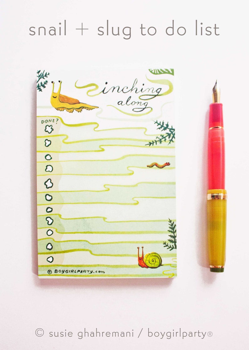 May include: A green and white to-do list notepad with a snail and slug illustration. The notepad has the text "inching along" and "DONE?" with a checklist. The notepad is on a white background with a red and yellow pen.