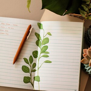 May include: An open notebook with lined pages and a brown pen. The notebook has a green leafy design on the right page. A succulent plant in a blue pot is on the right side of the notebook.