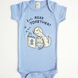 May include: A light blue baby onesie with a graphic of a dog and cat reading a book. The text on the onesie says "Let's Read Together!"
