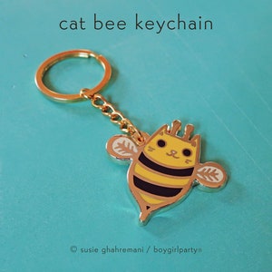May include: Gold-colored keychain featuring a cartoon cat bee with black stripes and yellow and white wings. The keychain has a gold-colored ring.