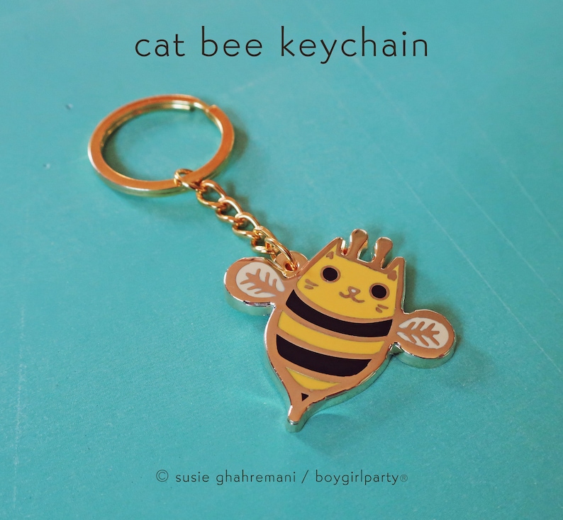May include: Gold-colored keychain featuring a cartoon cat bee with black stripes and yellow and white wings. The keychain has a gold-colored ring.