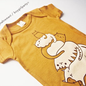 May include: A mustard yellow baby onesie with a graphic print of four white cats stacked on top of each other. The cats are drawn in a simple, cartoon style.