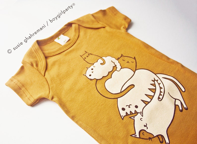 May include: A mustard yellow baby onesie with a graphic print of four white cats stacked on top of each other. The cats are drawn in a simple, cartoon style.