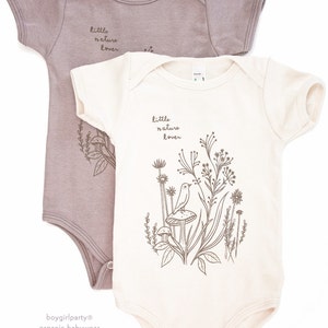 Puede incluir: Dos bodies para beb&eacute;, uno de color marr&oacute;n claro y otro de color crema. Ambos bodies tienen el texto "little nature lover" impreso en ellos. El body crema tiene un gr&aacute;fico de un p&aacute;jaro en un hongo con plantas y flores.