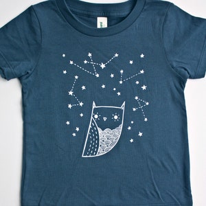 May include: A dark blue organic toddler tee with a white owl and star design. The owl is looking up at the stars, which are connected by lines. The text "susie ghahremani / boygirlparty&reg;" is at the bottom of the tee.