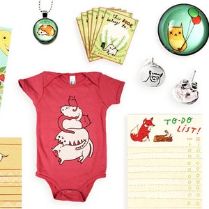 May include: A collection of items featuring cute animal illustrations.  The items include a book titled "What Will Hatch?", a red baby onesie with a stack of cats, a notepad with a fox and the words "To-Do List!" written on it, a small notebook with a woodland scene, and several small glass ornaments with animal designs.