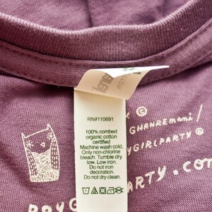 May include: A purple t-shirt with a white owl graphic and care instructions. The text on the tag reads "100% combed organic cotton certified" and "Machine wash cold, Only non-chlorine bleach. Tumble dry low. Low iron. Do not iron decoration. Do not dry clean."