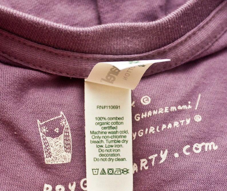 May include: A purple t-shirt with a white owl graphic and care instructions. The text on the tag reads "100% combed organic cotton certified" and "Machine wash cold, Only non-chlorine bleach. Tumble dry low. Low iron. Do not iron decoration. Do not dry clean."