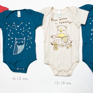 May include: Four baby onesies in different colors and sizes. The onesies feature various designs, including a stack of cats, a constellation with an owl, a bear reading a book with the text "BOOK WORM in TRAINing!" and a constellation with an owl. The onesies are labeled with the sizes 3-6 mo, 6-12 mo, 12-18 mo, and 18-24 mo.