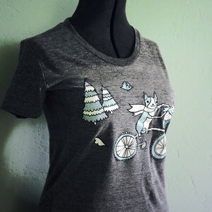 May include: A dark gray t-shirt with a graphic of a dog riding a bicycle with two trees and a bird in the background.