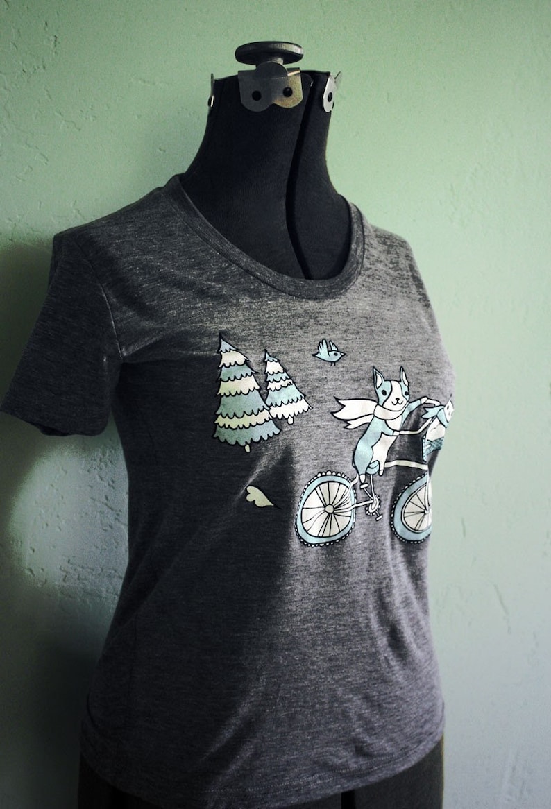May include: A dark gray t-shirt with a graphic of a dog riding a bicycle with two trees and a bird in the background.