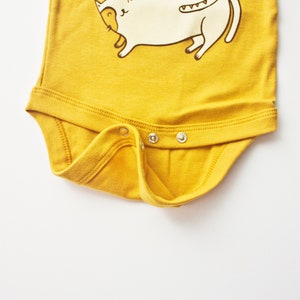 May include: A yellow baby onesie with a white cat illustration. The onesie has three snaps on the bottom.