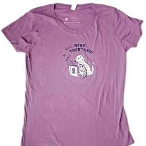 May include: Purple organic cotton t-shirt featuring a graphic of a dog reading a book to a cat. The text on the shirt reads "Let's Read Together!"