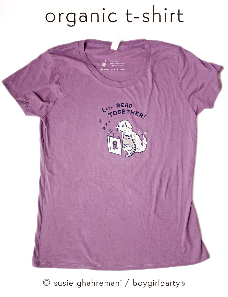 May include: Purple organic cotton t-shirt featuring a graphic of a dog reading a book to a cat. The text on the shirt reads "Let's Read Together!"