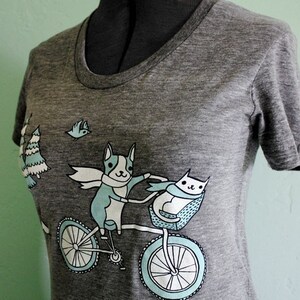 May include: A gray heather t-shirt with a graphic of a dog and cat riding a bicycle. The dog is wearing a scarf and the cat is in a basket. There are trees and a bird in the background.