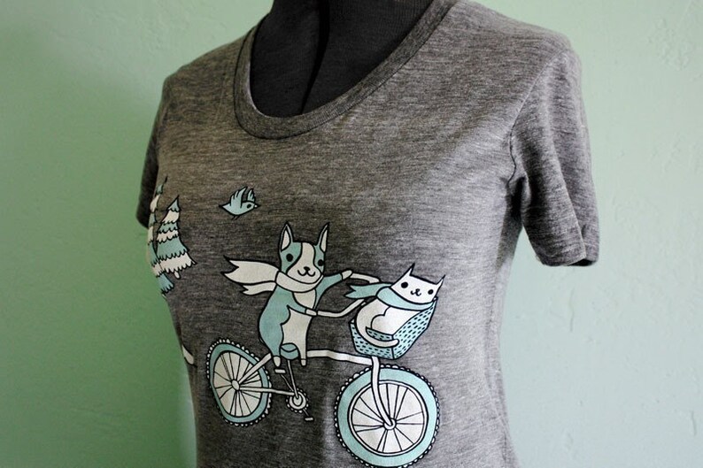 May include: A gray heather t-shirt with a graphic of a dog and cat riding a bicycle. The dog is wearing a scarf and the cat is in a basket. There are trees and a bird in the background.
