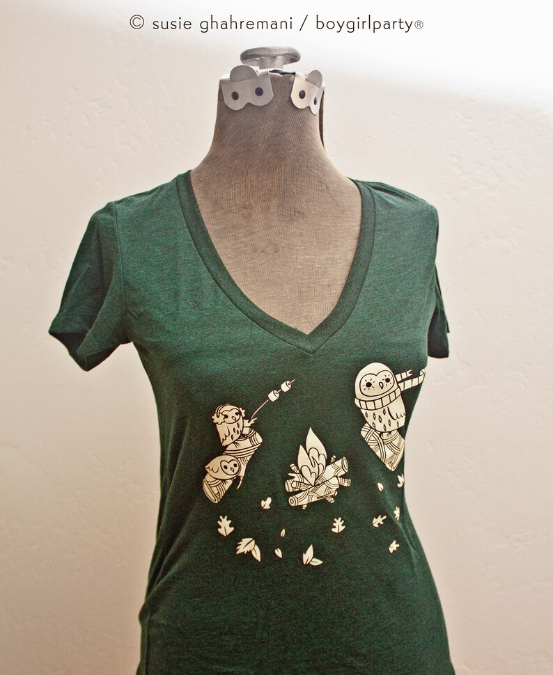 May include: A dark green v-neck t-shirt with a white graphic of three owls around a campfire. The owls are wearing hats and scarves. The graphic also includes leaves and a marshmallow on a stick.