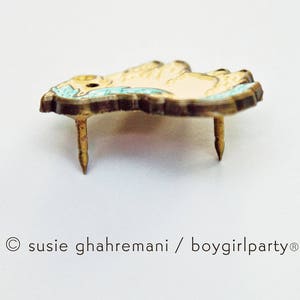 Op de afbeelding: Een gouden pin met een lichtbruin en blauw ontwerp. De pin is gevormd als een klein dier met een gekrulde staart en een puntige neus. De pin heeft twee metalen pinnen aan de achterkant.