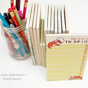 K&ouml;nnte beinhalten: Ein gelber Notizblock f&uuml;r To-Do-Listen mit einer roten Katzenillustration und dem Text "like it or not TO DO LIST" auf der Vorderseite. Der Notizblock liegt auf einem Tisch mit einem Glasgef&auml;&szlig; voller bunter Stifte und F&uuml;ller.