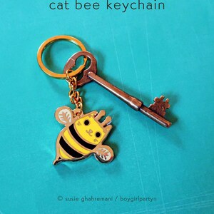 May include: A gold key chain with a small, yellow and black bee charm with a cat face. The bee charm has a gold outline and black stripes. The key chain is attached to a gold ring.