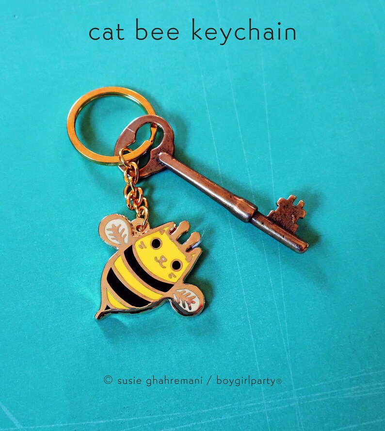 May include: A gold key chain with a small, yellow and black bee charm with a cat face. The bee charm has a gold outline and black stripes. The key chain is attached to a gold ring.