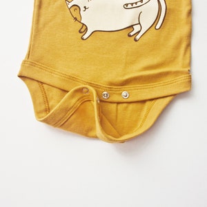 May include: A mustard yellow baby onesie with a white cartoon cat illustration. The onesie has three white snaps on the bottom.