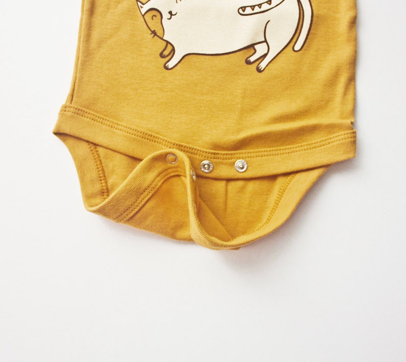 May include: A mustard yellow baby onesie with a white cartoon cat illustration. The onesie has three white snaps on the bottom.