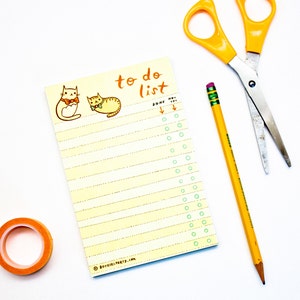 May include: A yellow to-do list notepad featuring two cartoon cats wearing bow ties. The notepad has a column of circles for ticking off tasks. The text "to do list" is written in orange at the top of the notepad. A yellow pencil and a pair of silver scissors are also in the image.