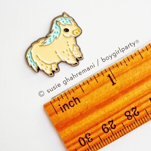 Op de afbeelding: Een kleine, gele en blauwe emaille pin van een pluche pony. De pin is ongeveer 2,5 cm hoog en ligt naast een liniaal ter referentie van de grootte.
