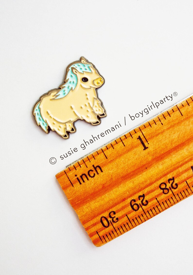 Op de afbeelding: Een kleine, gele en blauwe emaille pin van een pluche pony. De pin is ongeveer 2,5 cm hoog en ligt naast een liniaal ter referentie van de grootte.