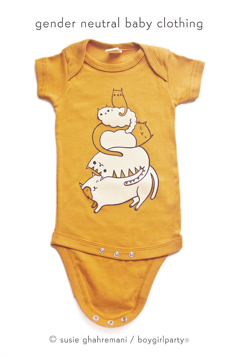 May include: A mustard yellow baby onesie with a graphic of a stack of four white cats with black eyes and brown outlines. The text "susie ghahremani / boygirlparty&reg;" is at the bottom of the onesie.