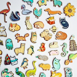 May include: A collection of colorful enamel pins featuring various animals, objects, and characters. The pins are arranged in a grid pattern on a white background. The text "enamel pins" and "boygirlparty.com" are visible in the image.