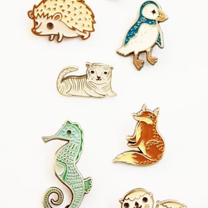 May include: A set of enamel pins featuring various animals, including a cat, hedgehog, puffin, tiger, fox, seahorse, and two otters. The pins are all in a cartoon style and have a gold finish. The text "boygirlparty&reg; pin collection" is at the top of the image.