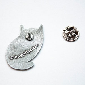 May include: Silver metal pin of a cat sitting down. The back of the pin has the text "&copy; boygirlparty&reg;" printed on it.