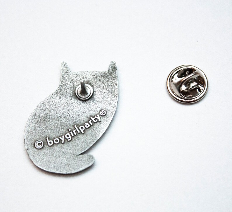 May include: Silver metal pin of a cat sitting down. The back of the pin has the text "&copy; boygirlparty&reg;" printed on it.