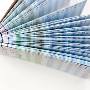 May include: A partially fanned-out booklet with pages displaying a repeating pattern of blue, green, and purple rectangles. The booklet's spine is visible, and the pages are arranged in a fan shape, creating a visual display.