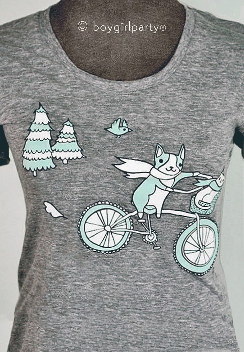 May include: A gray t-shirt with a graphic of a dog riding a bicycle through a forest. The dog is wearing a scarf and there are two trees and a bird in the background. The text "boygirlparty&reg;" is printed on the shirt.