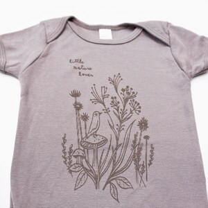 Puede incluir: Un body de beb&eacute; gris claro con un gr&aacute;fico marr&oacute;n de un p&aacute;jaro, hongos y flores. El texto "little nature lover" est&aacute; impreso en el body.