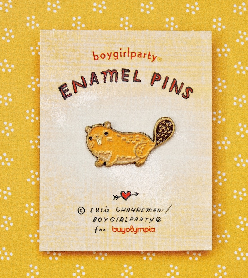 May include: A gold enamel pin of a yellow beaver with a red mouth and a brown tail. The pin is on a white card with the text 'boygirlparty® ENAMEL PINS' in black and red. The card also has the text '© susie GHAHRREMANI / BOYGIRLPARTY® for buyolympia' in black.
