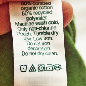 May include: White care label with green text and symbols. The label states "50% combed organic cotton 50% recycled polyester. Machine wash cold, Only non-chlorine bleach. Tumble dry low. Low iron. Do not iron decoration. Do not dry clean." The label also includes four symbols: a tub of water with a wavy line, a triangle with a circle inside, a circle with a dot inside, and a circle with a diagonal line through it. The label concludes with "Made in USA with imported yarns."
